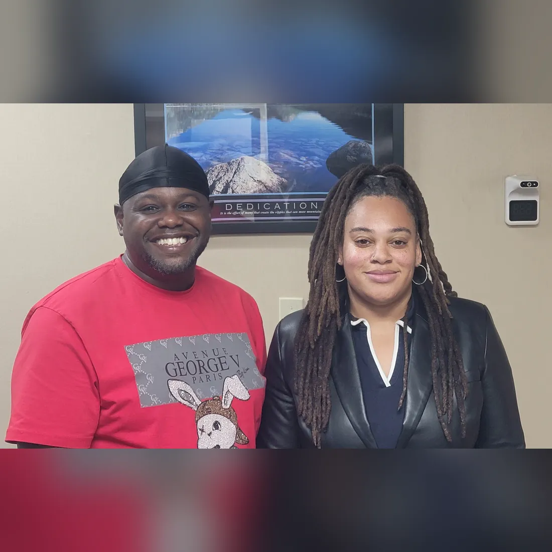 Two people smile together in front of a framed photo in an office